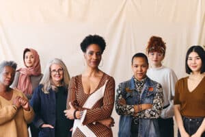 Group of diverse women facing the camera in front of a cream colored draped cloth. They look fierce and strong and proud.